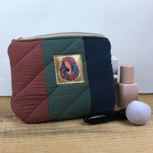 Könnte beinhalten: Eine gesteppte Kosmetiktasche mit Reißverschluss, mit einem Farbblock-Design in Burgunder-, Grün- und Marineblautönen. Auf der Vorderseite befindet sich ein goldfarbener quadratischer Aufnäher mit einer floralen Illustration einer Frau. Ein schwarzer Eyeliner und ein weißer Make-up-Schwamm liegen neben der Tasche.