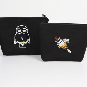 Könnte beinhalten: Zwei schwarze Reißverschlusstaschen mit gestickten Designs. Eine Tasche zeigt eine weiße Eule mit gelben Augen. Die andere Tasche zeigt eine weiße Flasche mit einer gelben Flüssigkeit und einem Etikett mit der Aufschrift "Take Potion".