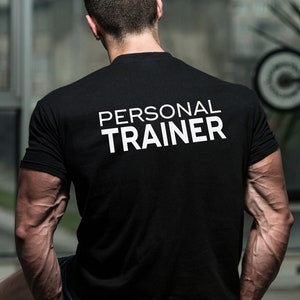 Pode incluir: Um homem vestindo uma camiseta preta com o texto "PERSONAL TRAINER" impresso em branco nas costas. Ele está em uma academia com equipamentos de musculação no fundo.