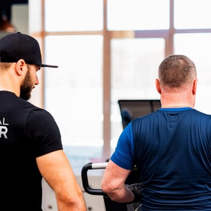 K&ouml;nnte beinhalten: Ein Personal Trainer in einem schwarzen Shirt mit dem Text "PERSONAL TRAINER" auf dem R&uuml;cken, der mit einem Kunden in einem blauen Shirt spricht. Der Kunde befindet sich auf einem Laufband.