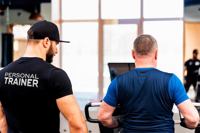 K&ouml;nnte beinhalten: Ein Personal Trainer in einem schwarzen Shirt mit dem Text "PERSONAL TRAINER" auf dem R&uuml;cken, der mit einem Kunden in einem blauen Shirt spricht. Der Kunde befindet sich auf einem Laufband.