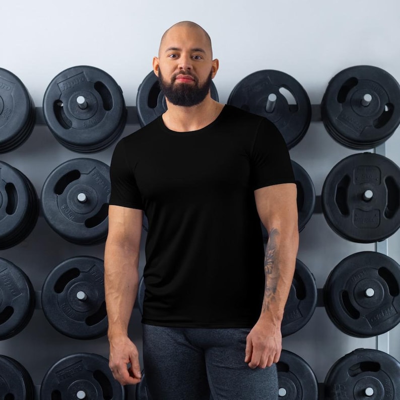 K&ouml;nnte beinhalten: Ein Mann in einem schwarzen T-Shirt und grauen Hosen steht vor einer Wand mit Gewichtsplatten.