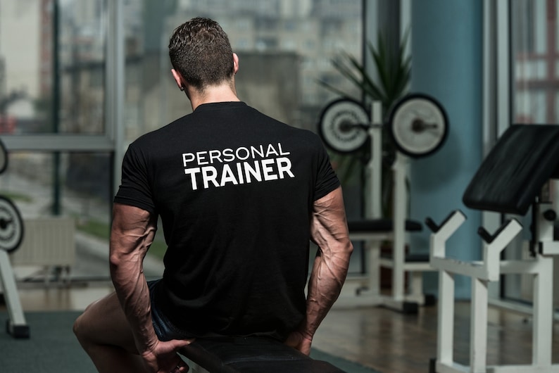 K&ouml;nnte beinhalten: Ein Mann tr&auml;gt ein schwarzes T-Shirt mit dem Text "PERSONAL TRAINER" in wei&szlig; auf dem R&uuml;cken. Er steht in einem Fitnessstudio mit Krafttrainingsger&auml;ten im Hintergrund.