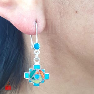 May include: A silver earring with a turquoise stone and a cross-shaped pendant. The pendant features a colorful geometric design with green, orange, and yellow accents. The earring is hanging from an ear.