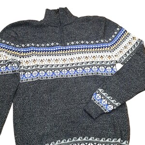 May include: A charcoal gray zip-up sweater with a patterned design in blue, white, and tan. The sweater features a high collar and ribbed cuffs. The pattern includes geometric shapes and stylized animal figures.