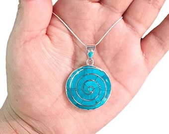 Può includere: Una collana d'argento con un ciondolo rotondo con un design a spirale turchese. Il ciondolo è composto da piccoli pezzi di turchese incastonati in una cornice d'argento. La collana è tenuta in una mano.