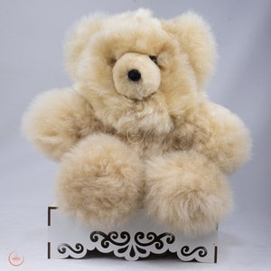 May include: A beige alpaca teddy bear sitting on a white decorative stand. The bear has a fluffy, soft texture and a sweet expression.
