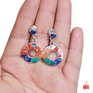 May include: A pair of silver earrings with a round, open-circle design. The earrings are made with a variety of colorful stones, including blue, green, orange, and white. The earrings are hanging from a small blue stone.