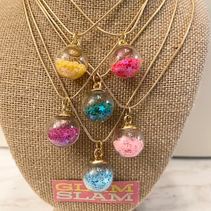 May include: Six gold chain necklaces with clear glass globe pendants filled with colorful glitter. Each pendant contains a different color of glitter, including pink, blue, green, yellow, and white. The necklaces are displayed on a brown burlap mannequin. The text "GLAM SLAM" is printed on a pink and white label below the necklaces.