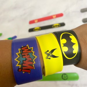 Peut inclure: Trois bracelets en caoutchouc colorés avec des motifs différents. Le premier bracelet est bleu avec une étoile jaune et le texte "WHAM!" Le deuxième bracelet est jaune avec un symbole noir. Le troisième bracelet est noir avec un logo Batman noir.