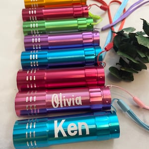 Może przedstawiać: Zestaw ośmiu kolorowych mini latarek z personalizowanymi nazwami, w tym "Olivia" i "Ken". Każda latarka ma sznurek do łatwego przenoszenia.