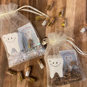 May include: Two white tooth-shaped wooden ornaments are in separate sheer white organza bags. Each bag contains a small card with the text "I lost a tooth! I am a special kid!" and a small glass vial filled with gold glitter. The bags are on a wooden surface.