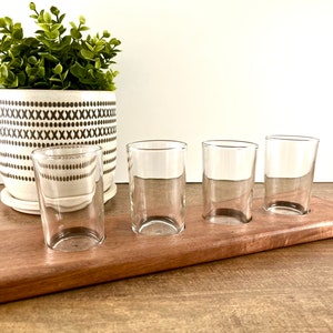 May include: A wooden serving tray with four clear glass shot glasses. The tray is made of dark wood and has a natural finish. The glasses are arranged in a row on the tray.