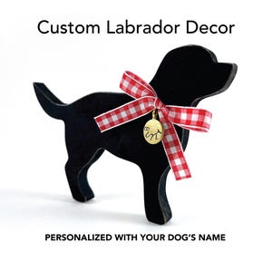 May include: Black Labrador silhouette with a red and white gingham bow and a gold charm. The text "Custom Labrador Decor" and "Personalized with your dog's name" are displayed above and below the dog silhouette.