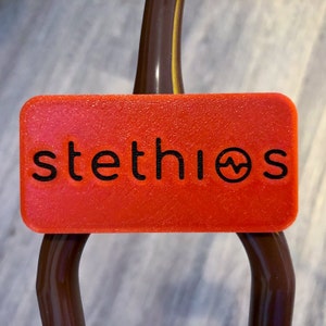 Puede incluir: Una etiqueta rectangular roja brillante para estetoscopio con la palabra "stethios" en letras negras. La etiqueta es texturizada y tiene un pequeño gráfico de una forma de onda. La etiqueta está colocada en un estetoscopio marrón.