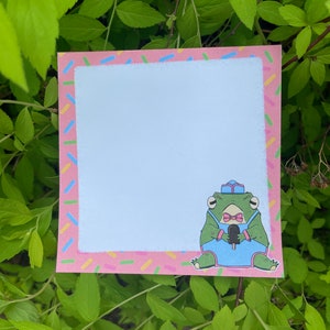Peut inclure: Un bloc-notes carré avec une bordure rose décorée de vermicelles colorés. Une grenouille de dessin animé portant un uniforme et un chapeau bleus tient une glace. Le bloc-notes est entouré de feuilles vertes.