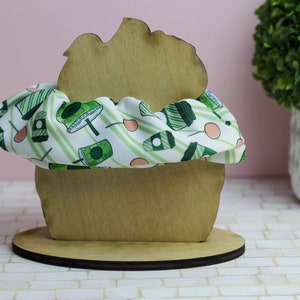 Op de afbeelding: Een houten cupcake-standaard met een groene en witte gestreepte scrunchie van stof erop. De standaard staat op een witte betegelde ondergrond.