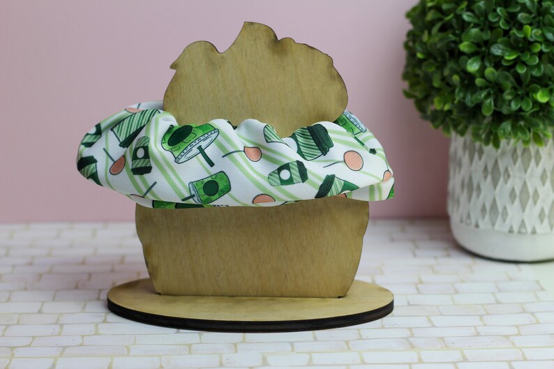 Op de afbeelding: Een houten cupcake-standaard met een groene en witte gestreepte scrunchie van stof erop. De standaard staat op een witte betegelde ondergrond.