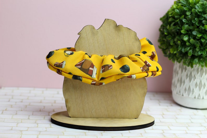 Op de afbeelding: Een houten haarbandhouder in de vorm van een hoofd met een gele haarband met bruine en zwarte ontwerpen.