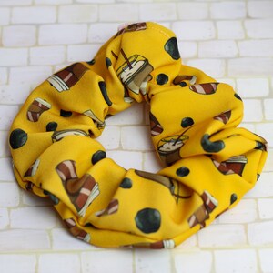 Op de afbeelding: Gele scrunchie met een patroon van bruine koffiekopjes en zwarte stippen.