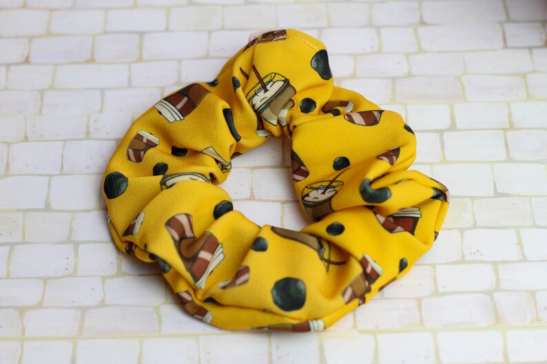 Op de afbeelding: Gele scrunchie met een patroon van bruine koffiekopjes en zwarte stippen.