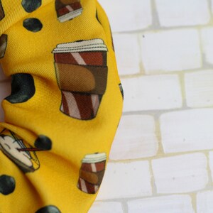 Op de afbeelding: Gele scrunchie met een herhalend patroon van koffiebekers en zwarte stippen.