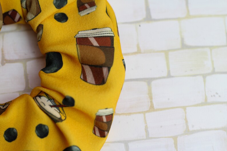Op de afbeelding: Gele scrunchie met een herhalend patroon van koffiebekers en zwarte stippen.