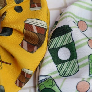 Op de afbeelding: Twee stoffen haarbandjes met een koffiekopjespatroon. Een haarbandje is geel met bruine koffiekopjes en de andere is wit met groene koffiekopjes.