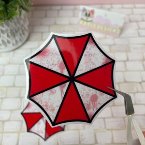 May include: A red and white umbrella sticker with a black outline and blood splatter design. The sticker is being peeled off of a white tiled surface with tweezers.