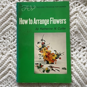 May include: A vintage book titled "How to Arrange Flowers" by Katherine N. Cutler. The cover is green with white text and a floral arrangement illustration. The book is part of the Amy Vanderbilt Success Program for Women.