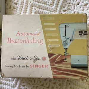 May include: Vintage Singer sewing machine box with the words "Automatic Buttonholing" in pink script. The box features an illustration of a sewing machine and the text "Touch & Sew". The box is cream and yellow.