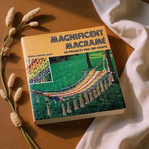May include: A book titled "Magnificent Macramé" with the subtitle "50 Projects You Can Create." The cover features a colorful macramé hammock design against a green background. The book is on a wooden surface with decorative wheat stalks.