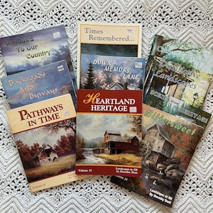 May include: A collection of vintage books with landscape paintings on the covers. Titles include "Windows To Our Country," "Backroads And Byways," "Pathways In Time," "Times Remembered," and "Heartland Heritage." The books are arranged on a white, textured surface.