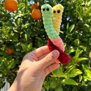 May include: Two crocheted worm plushies, one green and one yellow, are intertwined and held in a hand. The plushies have black eyes and are made with a fuzzy yarn.