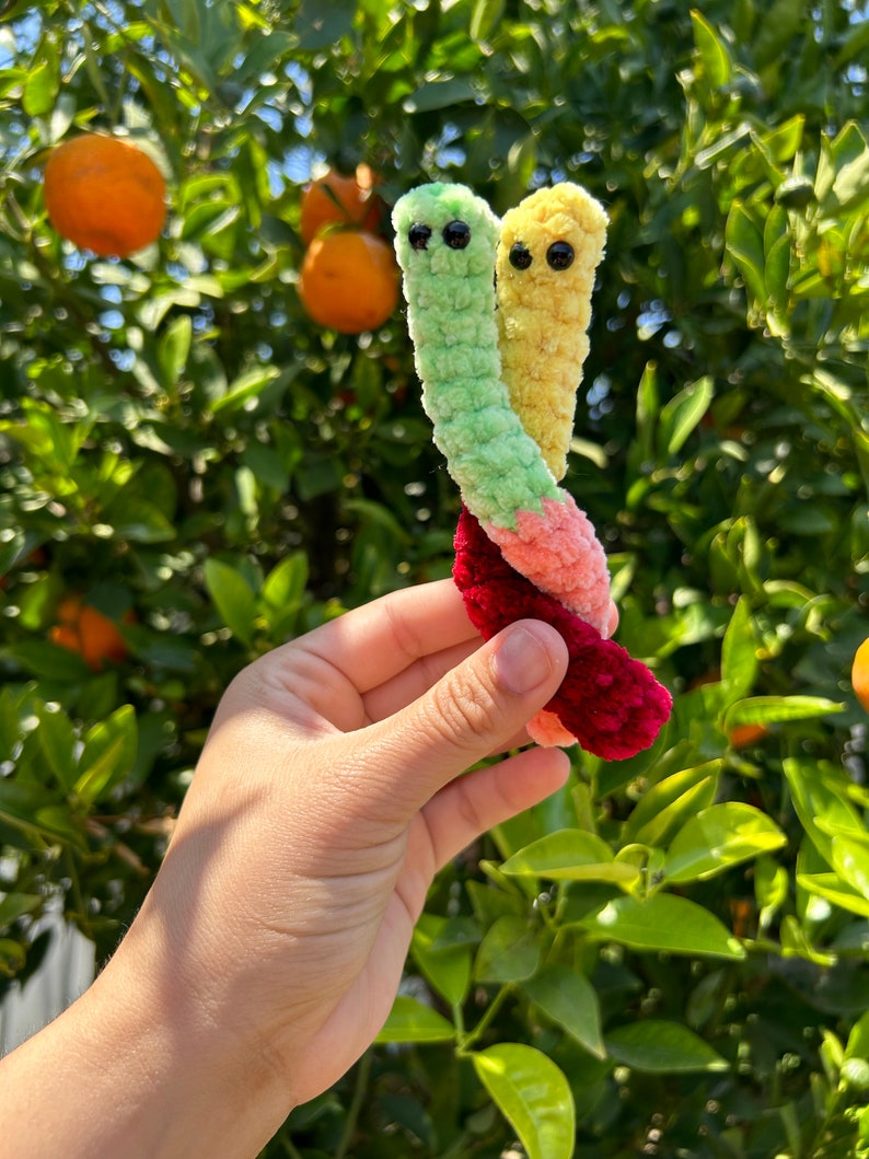May include: Two crocheted worm plushies, one green and one yellow, are intertwined and held in a hand. The plushies have black eyes and are made with a fuzzy yarn.