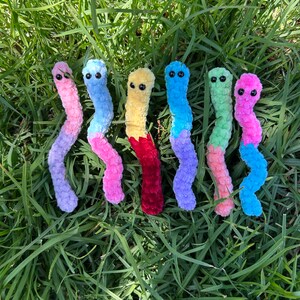 May include: Seven colorful crocheted worm plushies in various colors including pink, blue, yellow, red, green, and purple. The plushies are arranged in a row on a bed of green grass.