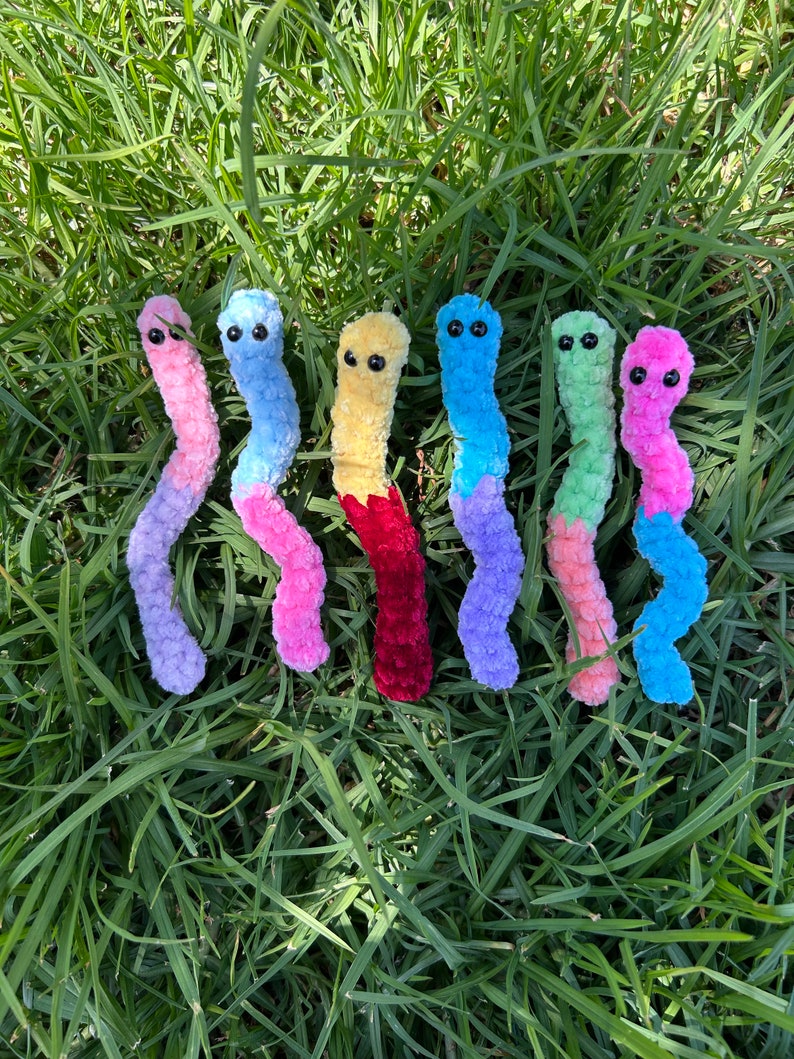 May include: Seven colorful crocheted worm plushies in various colors including pink, blue, yellow, red, green, and purple. The plushies are arranged in a row on a bed of green grass.