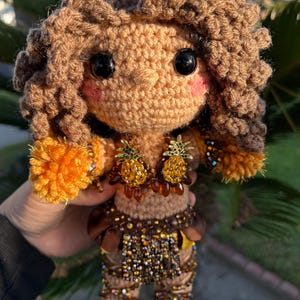 May include: A handcrafted crochet doll featuring curly brown yarn hair, large black eyes, and pink cheeks. The doll wears a top adorned with two beaded pineapple embellishments and a skirt decorated with brown fabric, beads, and sequins.