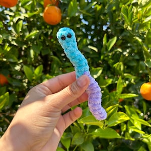 May include: A blue and purple crocheted worm with black eyes. The worm is being held by a hand in front of a green leafy background.