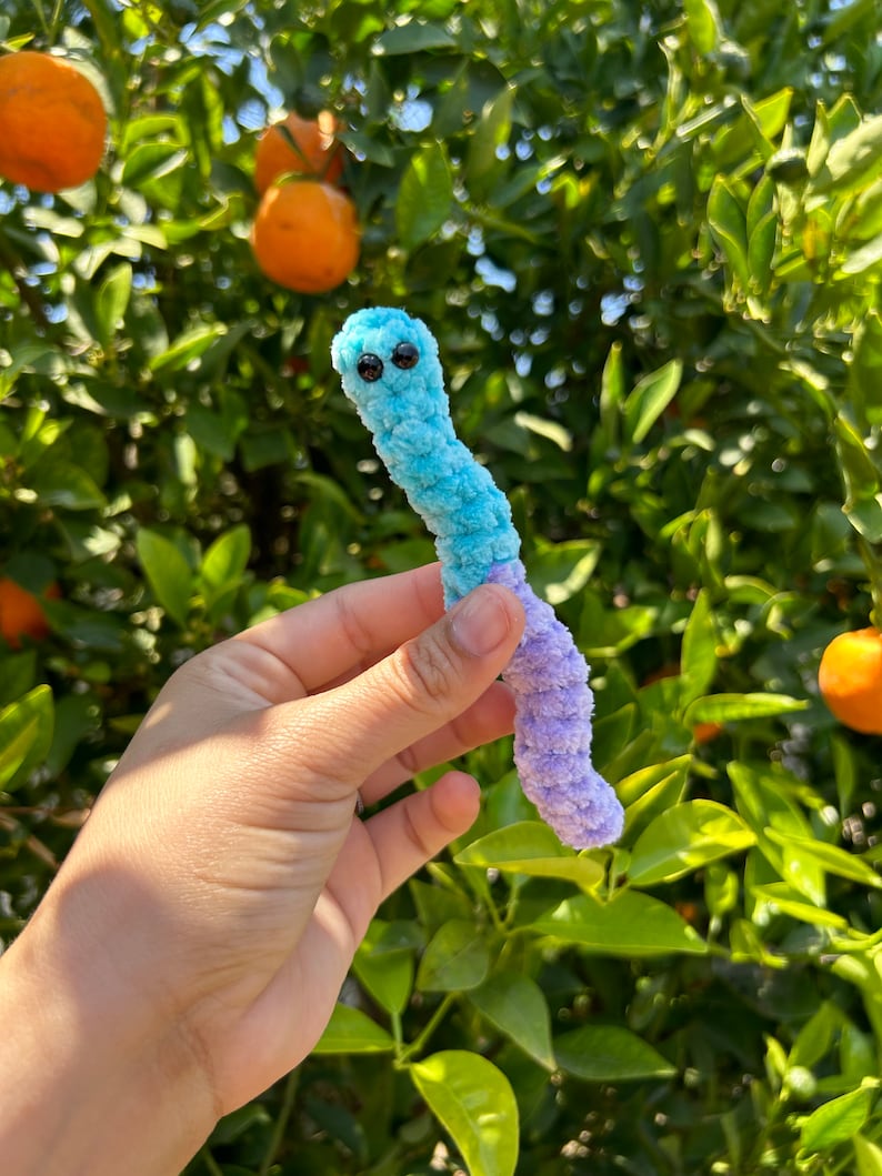 May include: A blue and purple crocheted worm with black eyes. The worm is being held by a hand in front of a green leafy background.