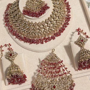 Puede incluir: Un conjunto de joyas de novia en tono dorado con collar, tocado y pendientes. El collar y el tocado tienen un dise&ntilde;o floral con detalles de cuentas rojas. Los pendientes tienen un dise&ntilde;o a juego. Las joyas se exhiben sobre una superficie color crema.