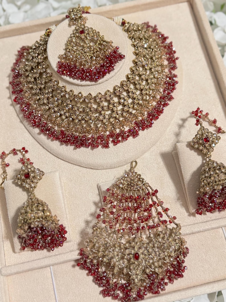 Puede incluir: Un conjunto de joyas de novia en tono dorado con collar, tocado y pendientes. El collar y el tocado tienen un dise&ntilde;o floral con detalles de cuentas rojas. Los pendientes tienen un dise&ntilde;o a juego. Las joyas se exhiben sobre una superficie color crema.