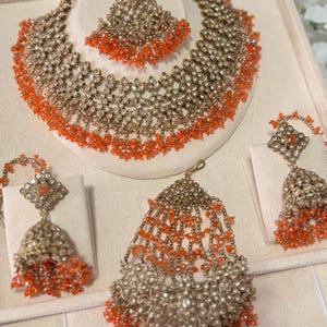 Puede incluir: Un conjunto de joyas ornamentado que incluye un collar, una pieza para la cabeza y pendientes. El conjunto es dorado con detalles intrincados y adornado con cuentas naranjas. El collar y la pieza para la cabeza est&aacute;n dise&ntilde;ados con un patr&oacute;n floral.