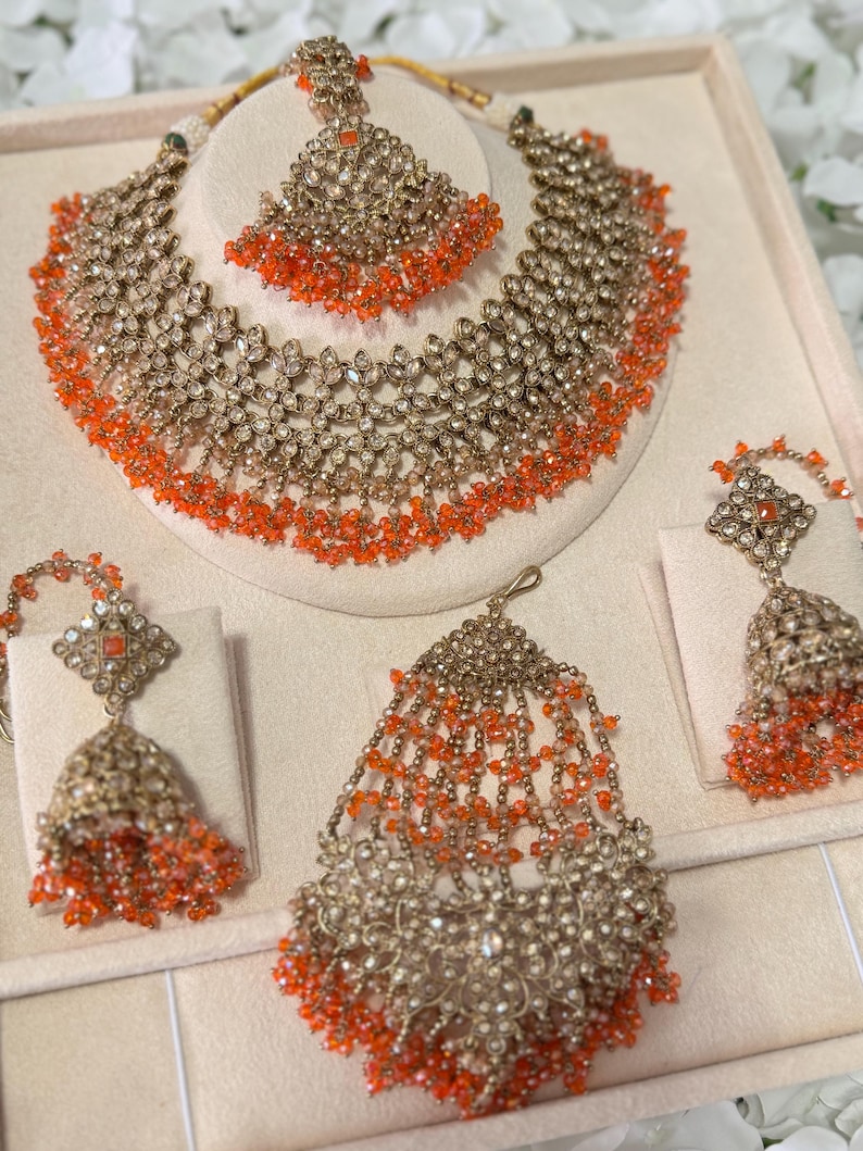 Puede incluir: Un conjunto de joyas ornamentado que incluye un collar, una pieza para la cabeza y pendientes. El conjunto es dorado con detalles intrincados y adornado con cuentas naranjas. El collar y la pieza para la cabeza est&aacute;n dise&ntilde;ados con un patr&oacute;n floral.
