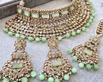 May include: A gold and green Indian bridal necklace set with a choker, earrings, and a maang tikka. The set features a layered design with green stones and pearls.