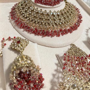 Puede incluir: Un conjunto de joyas dorado que incluye un collar, pendientes y una diadema. El conjunto est&aacute; adornado con cuentas de cristal rojas y transparentes. El collar y la diadema tienen un dise&ntilde;o floral. Los pendientes tienen un dise&ntilde;o colgante.