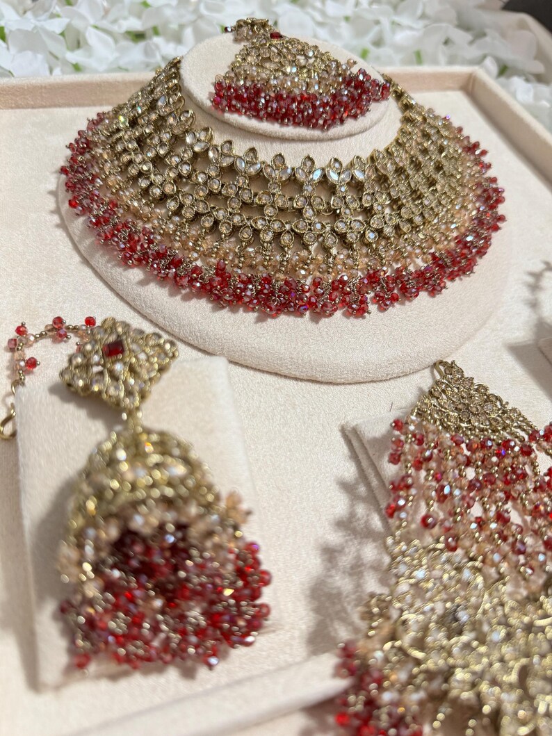 Puede incluir: Un conjunto de joyas dorado que incluye un collar, pendientes y una diadema. El conjunto est&aacute; adornado con cuentas de cristal rojas y transparentes. El collar y la diadema tienen un dise&ntilde;o floral. Los pendientes tienen un dise&ntilde;o colgante.