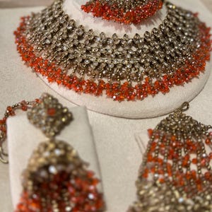 Puede incluir: Un conjunto de joyas de novia en tono dorado que incluye collar, pendientes y tocado. El collar y el tocado se exhiben en una cabeza de maniqu&iacute;. Las joyas est&aacute;n adornadas con cuentas transparentes y de color rojo anaranjado.
