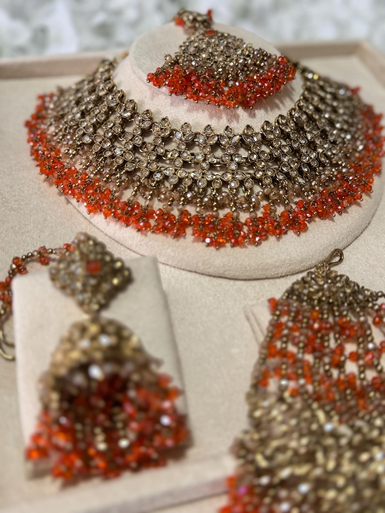 Puede incluir: Un conjunto de joyas de novia en tono dorado que incluye collar, pendientes y tocado. El collar y el tocado se exhiben en una cabeza de maniqu&iacute;. Las joyas est&aacute;n adornadas con cuentas transparentes y de color rojo anaranjado.