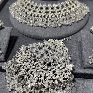 Puede incluir: Un deslumbrante conjunto de joyas de novia con tiara, collar y pulsera. El conjunto est&aacute; hecho de cristales transparentes y brillantes dispuestos en un intrincado dise&ntilde;o floral. Las joyas se exhiben sobre una superficie de terciopelo gris.